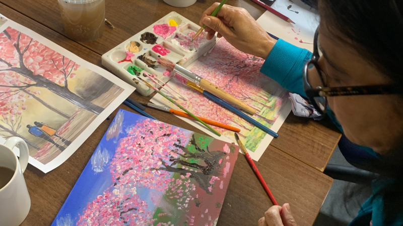 A person using a brush to mix pink paint on a palette while working on cherry blossom landscape paintings.