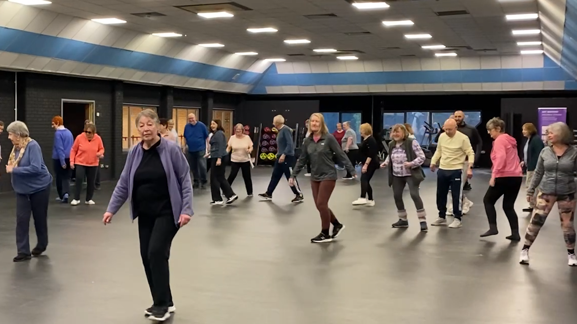 A group of older people wearing gym clothes participating in a fitness class in a large gym space.