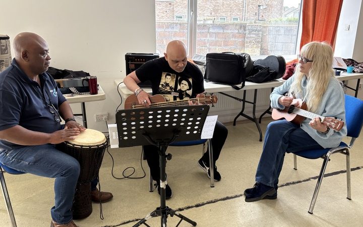 Three people sitting in a circle playing music together with a djembe drum, an acoustic bass, and a ukulele.