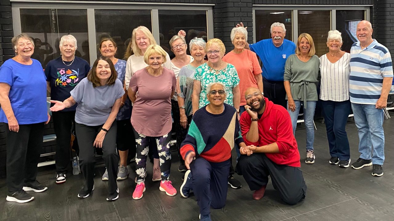 A large group of mostly older adults are posing and smiling for a group photo in a room.